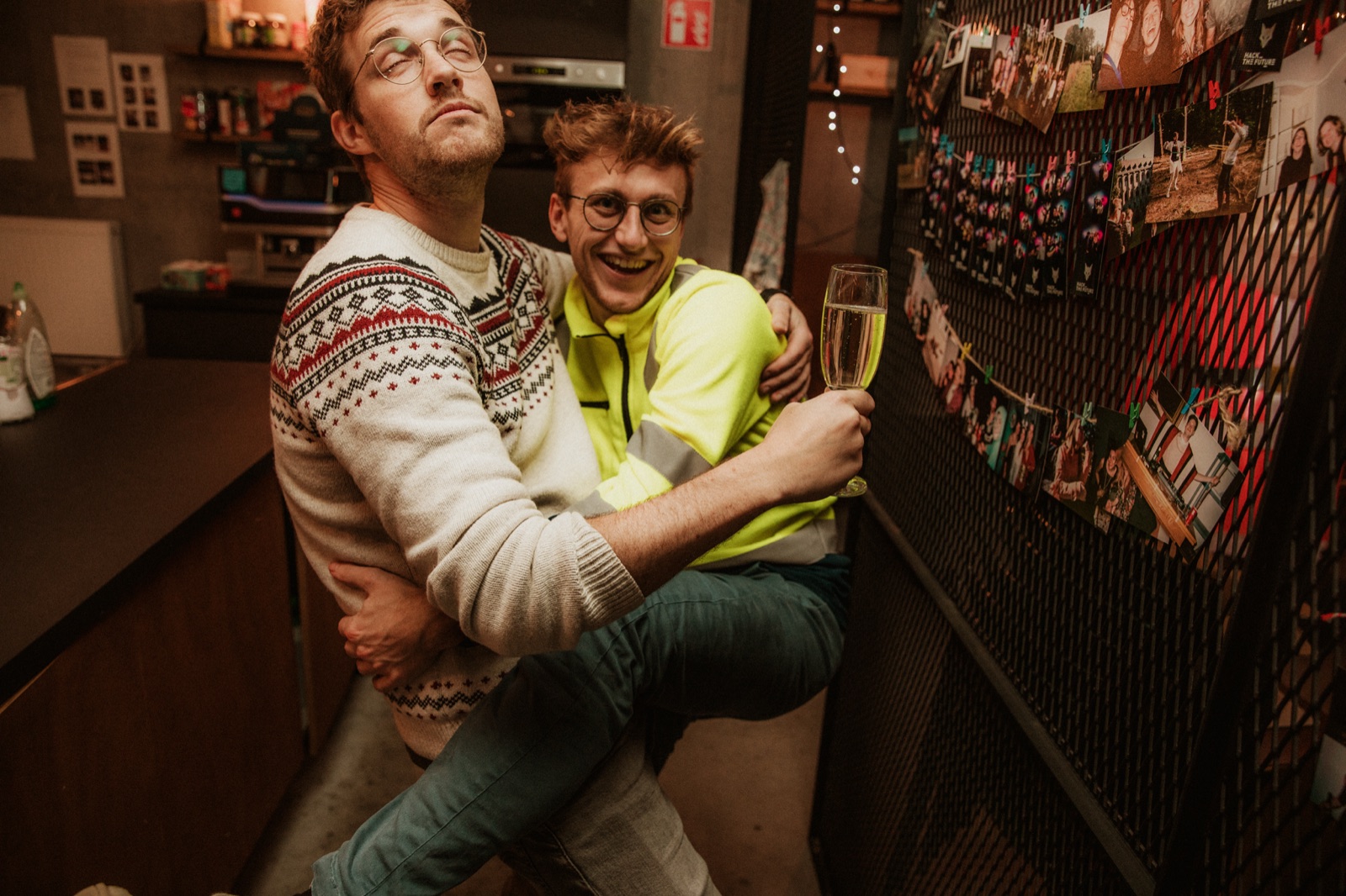 Two colleagues hugging and hamming it up with a glass of bubbles in the studio kitchen, next to a wall of pinned party photos